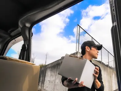 配達員が車から段ボール箱を取り出し、青空の下で運ぶ。商品発送・配送業務の実作業。