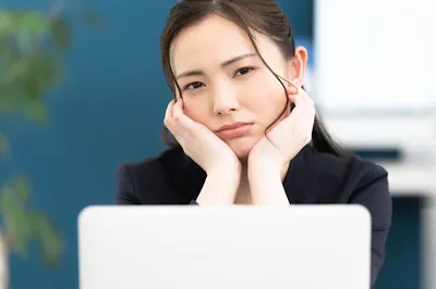 ノートパソコンの前で頭を抱えてストレス表情の黒いジャケット姿の女性。業務負担・システムの複雑さの課題表現