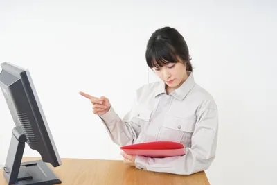 パソコン前で赤いフォルダを持ち指差す女性。倉庫・在庫業務の管理者