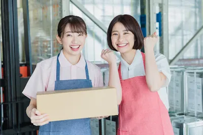 段ボール箱を持ち笑顔でポーズする2人の店舗スタッフ（青・ピンク色のエプロン）。EC業務・スタッフの満足度・チームワーク