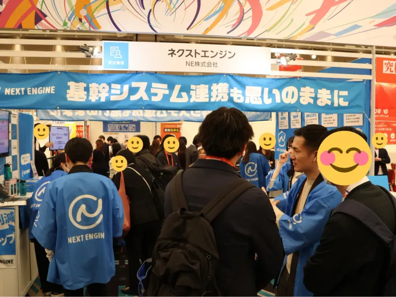 多くの来場者で賑わうネクストエンジンブースの様子。青い法被を着たスタッフがお客様に受注管理の説明をしているシーン。